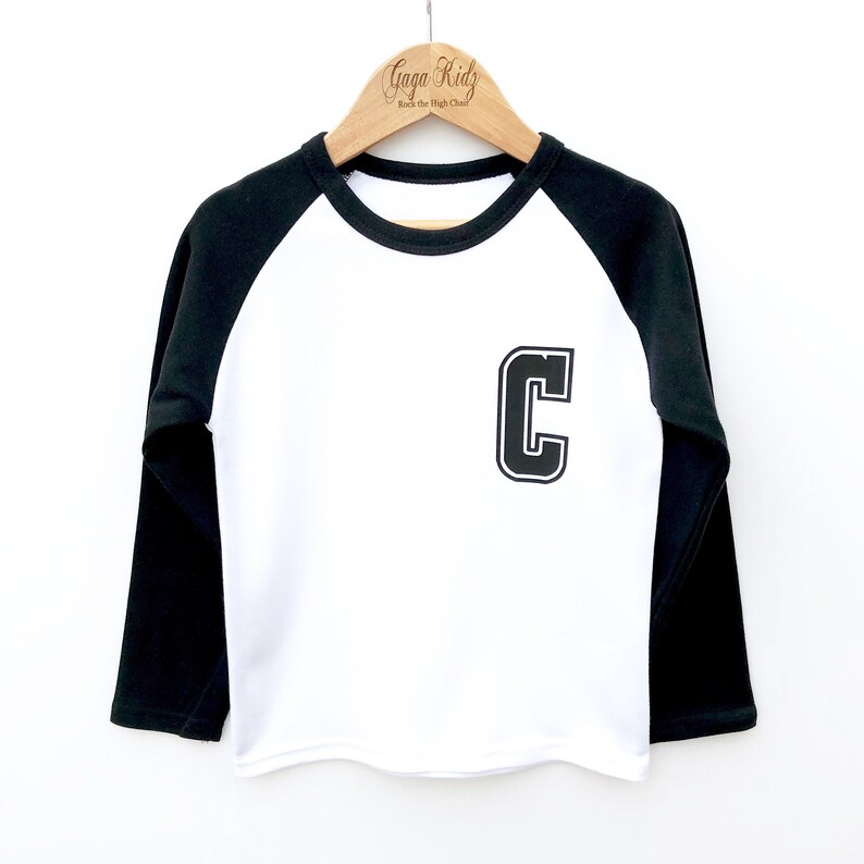 May include: White long-sleeved baseball tee with black sleeves and a black felt letter "C" on the front. The shirt is hanging on a wooden hanger with the text "Gaga Kidz Rock the High Chart" on it.
