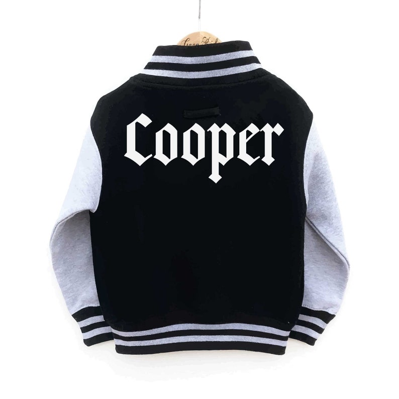 Op de afbeelding: Zwart en grijs varsity jack met witte letters op de rug die "Cooper" zeggen.