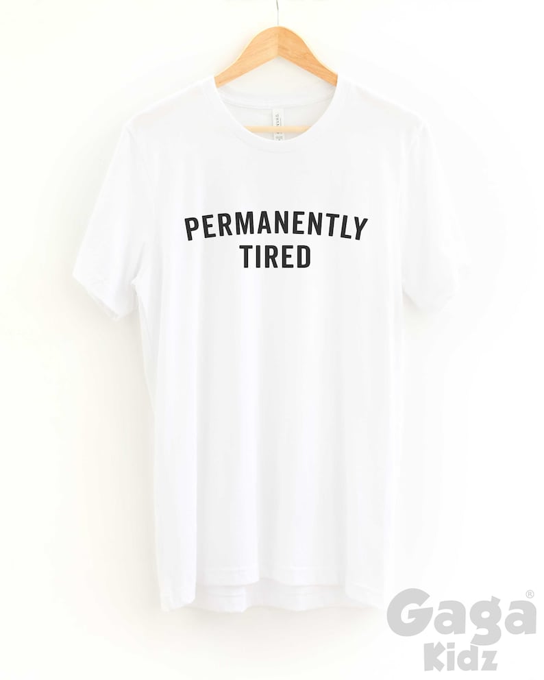 Puede incluir: Camiseta blanca con texto negro en may&uacute;sculas que dice "PERMANENTLY TIRED" en una percha.
