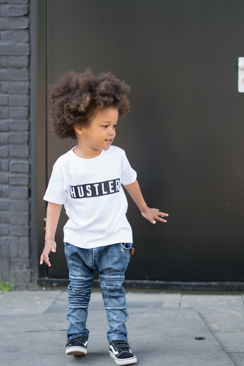 May include: A young boy with curly brown hair is wearing a white t-shirt with the word "Hustler" printed in black on it. He is also wearing blue jeans and black sneakers.