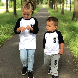 May include: Two children wearing matching white and black baseball shirts with the letters "M" and "S" on them. The girl is wearing blue jeans and the boy is wearing grey sweatpants. They are walking down a path lined with trees.