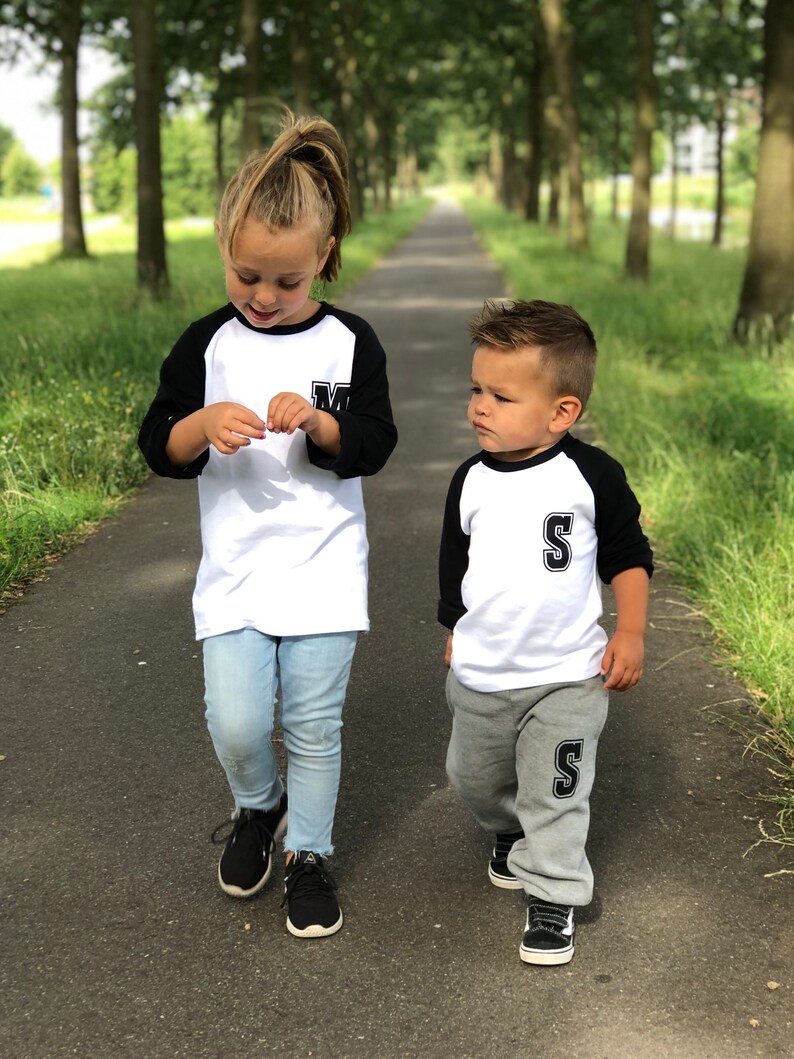 May include: Two children wearing matching white and black baseball shirts with the letters "M" and "S" on them. The girl is wearing blue jeans and the boy is wearing grey sweatpants. They are walking down a path lined with trees.