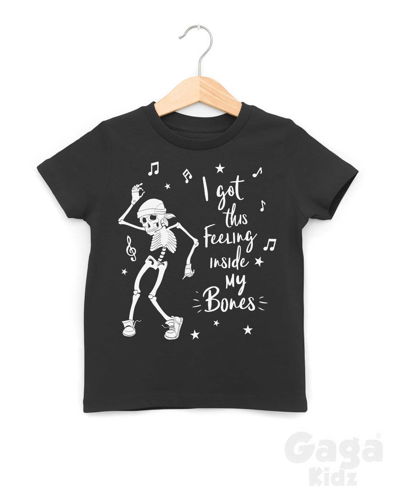 Peut inclure: T-shirt noir avec un squelette blanc dansant et le texte "I got this feeling inside my bones" avec des notes de musique et des &eacute;toiles.