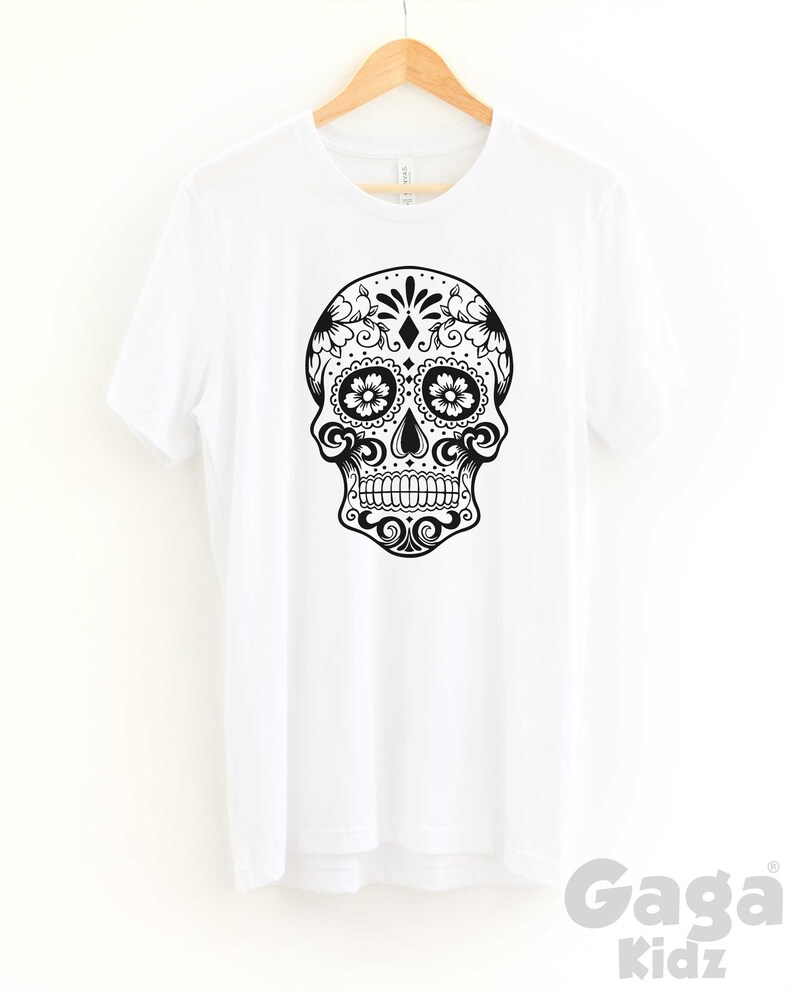 May include: White t-shirt with a black and white sugar skull graphic. The skull is decorated with floral patterns and has a diamond in the center of the forehead.