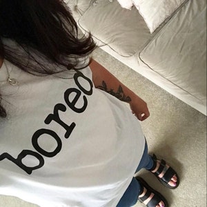 May include: A white t-shirt with the word "bored" printed in black. The person is wearing blue jeans and black sandals.