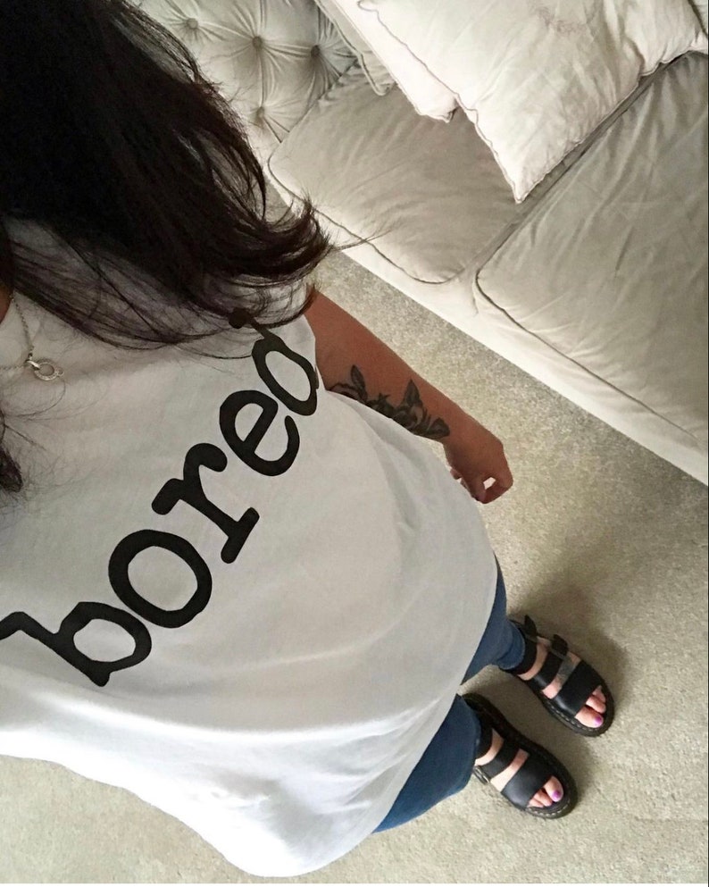 May include: A white t-shirt with the word "bored" printed in black. The person is wearing blue jeans and black sandals.