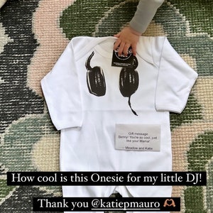 May include: A white long-sleeve onesie featuring a black graphic print of headphones on the chest. A small rectangular card is placed on the garment with the text: Gift message Benny! You're so cool, just like your Mama! -- Meadow and Katie.