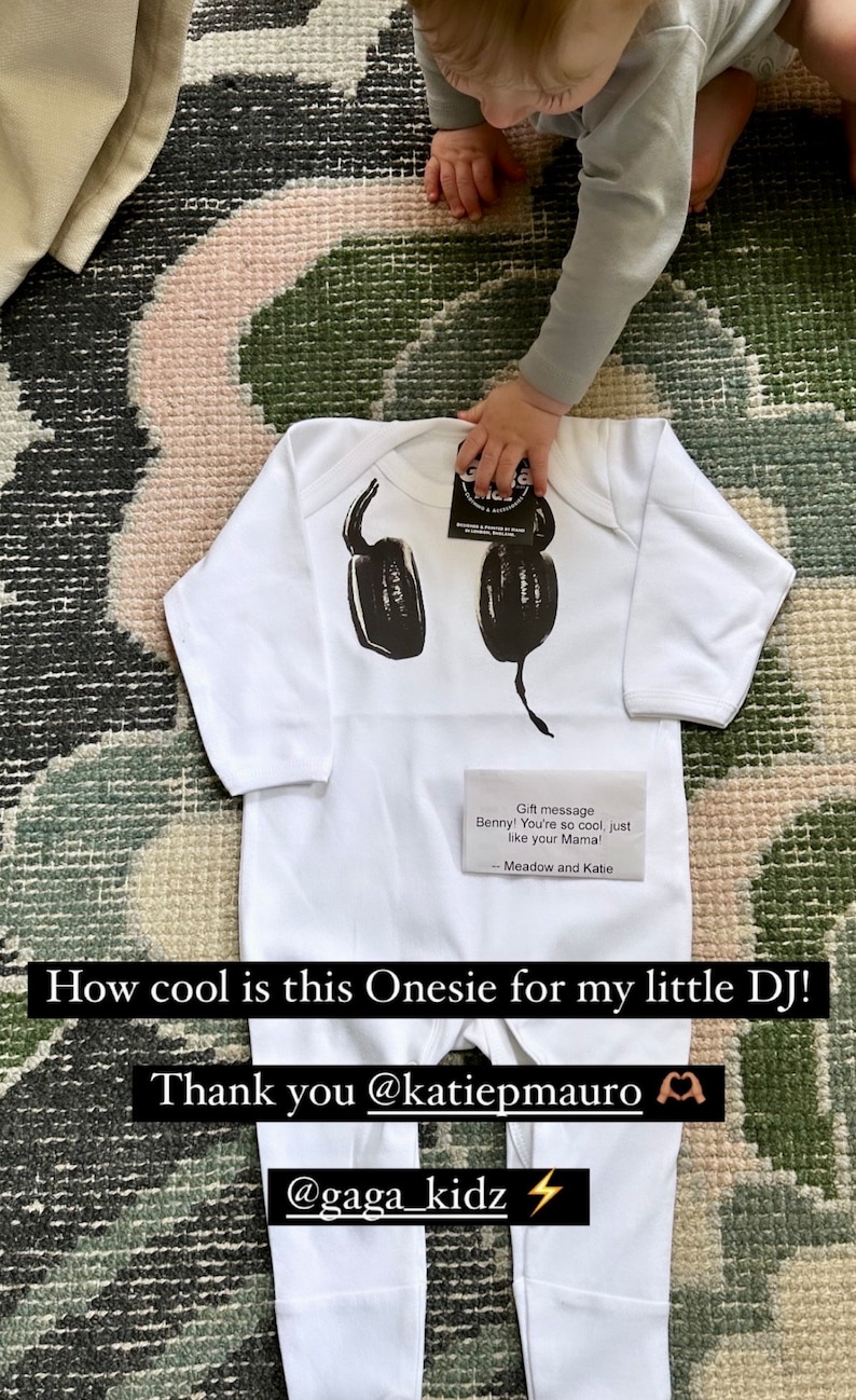 May include: A white long-sleeve onesie featuring a black graphic print of headphones on the chest. A small rectangular card is placed on the garment with the text: Gift message Benny! You're so cool, just like your Mama! -- Meadow and Katie.