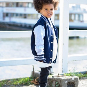 Op de afbeelding: Een jonge jongen draagt een donkerblauw en grijs baseballjack met witte strepen op de mouwen. Hij draagt een zwarte jeans en zwart-witte sneakers. Hij staat voor een wit hek.