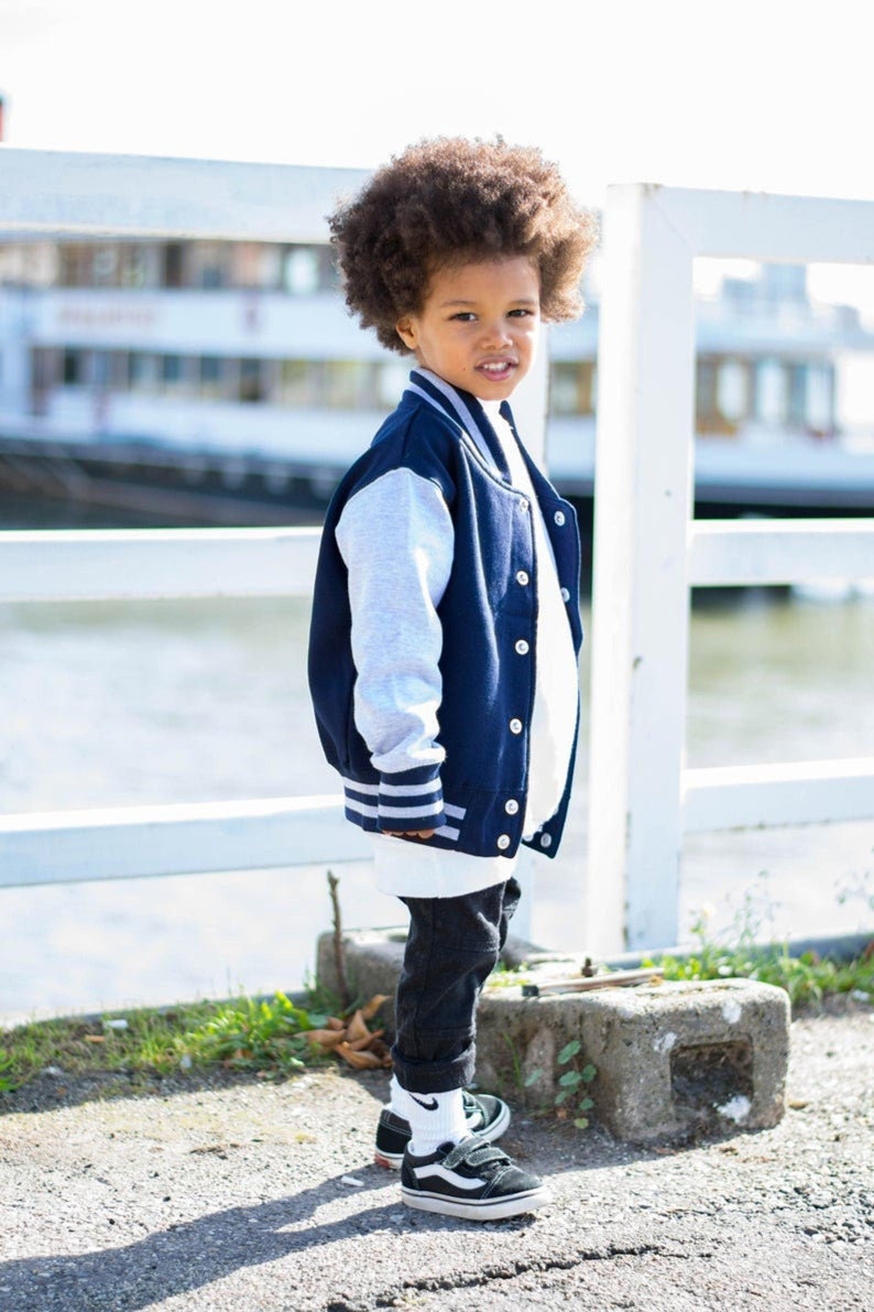 Op de afbeelding: Een jonge jongen draagt een donkerblauw en grijs baseballjack met witte strepen op de mouwen. Hij draagt een zwarte jeans en zwart-witte sneakers. Hij staat voor een wit hek.
