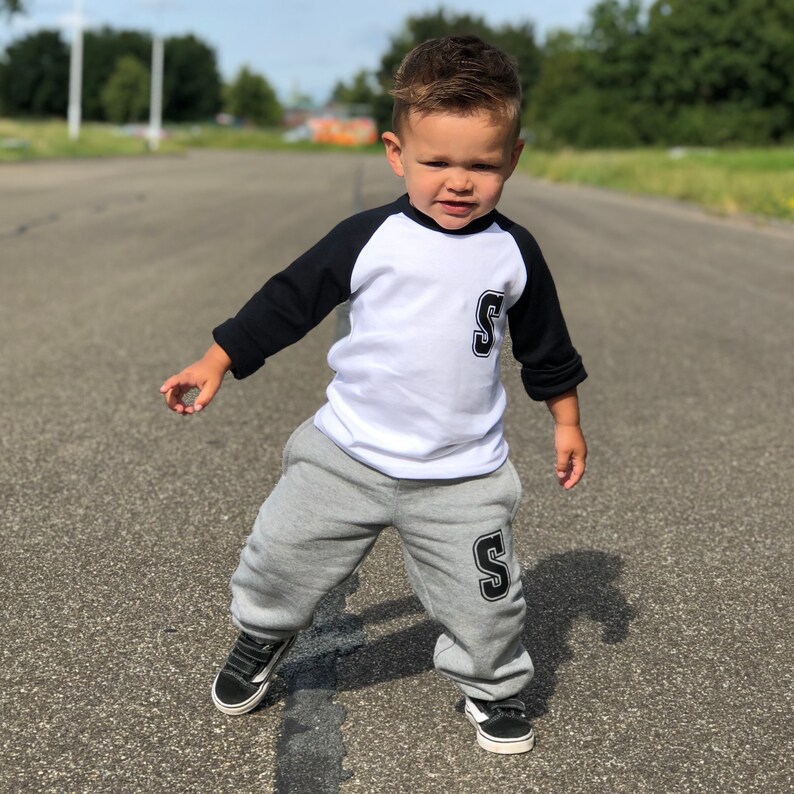 May include: A young child wearing a white and black baseball tee with a large black letter 'S' on the front and matching grey sweatpants with a black letter 'S' on the leg. The child is standing on a paved road.