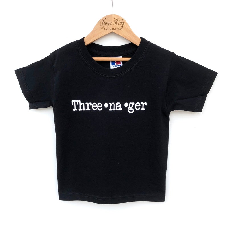 K&ouml;nnte beinhalten: Schwarzes T-Shirt mit wei&szlig;em Text "Three-na-ger" auf einem Holzb&uuml;gel.