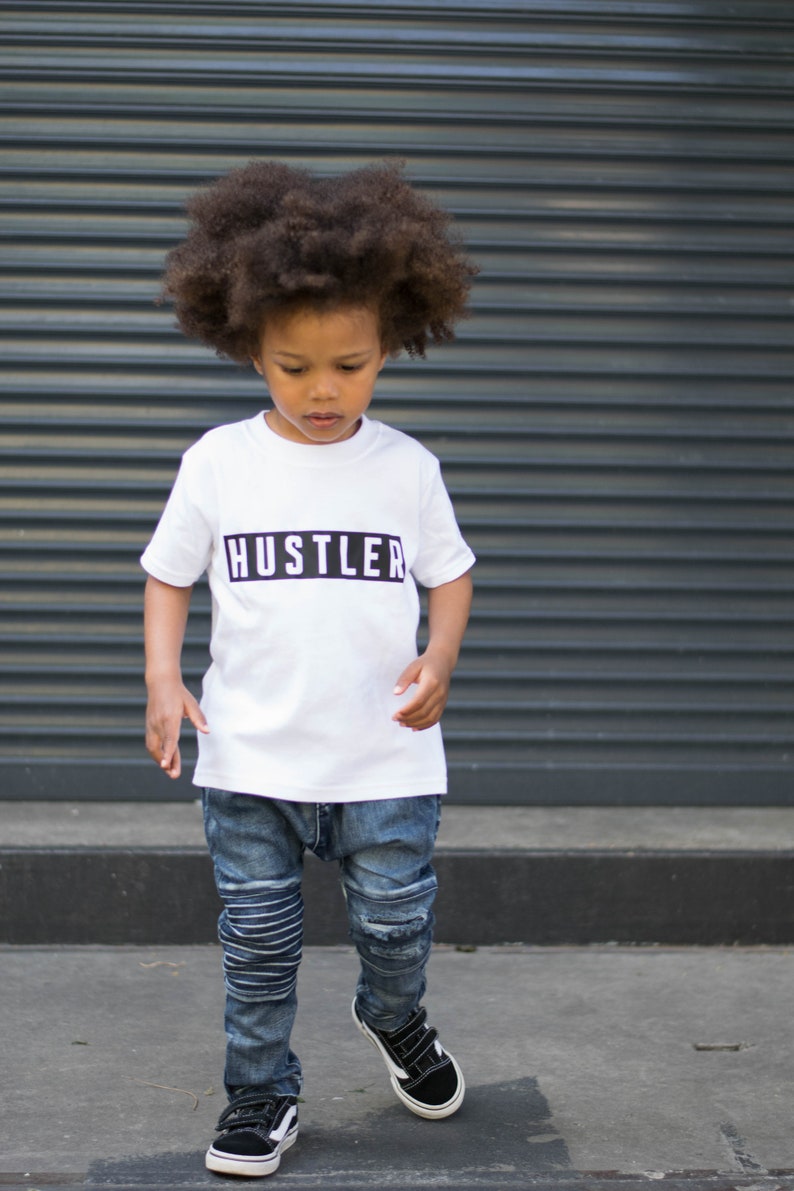 May include: A young person wearing a white t-shirt with the word "Hustler" printed in black on it. They are wearing blue jeans and black sneakers.