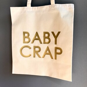 Pode incluir: Uma bolsa de lona branca com as palavras "BABY CRAP" impressas em dourado.