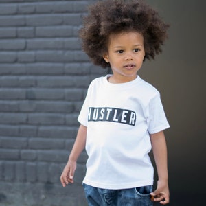 May include: A young person wearing a white t-shirt with the word "Hustler" printed in black on a black rectangle. The person has curly dark brown hair and is wearing blue jeans.
