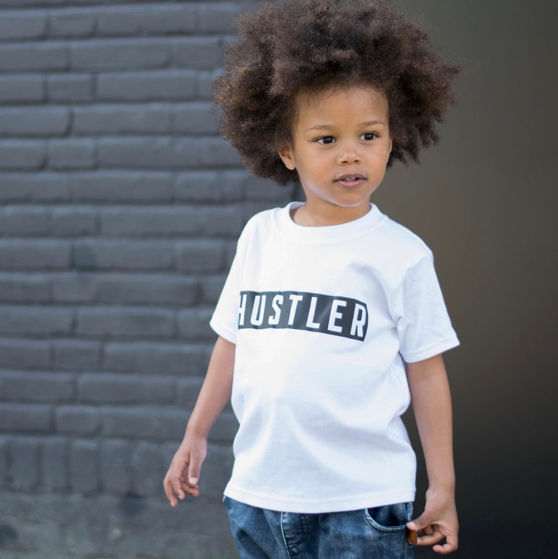 May include: A young person wearing a white t-shirt with the word "Hustler" printed in black on a black rectangle. The person has curly dark brown hair and is wearing blue jeans.