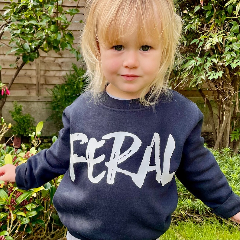 May include: A black sweatshirt with the word "FERAL" written in white paint across the front. The sweatshirt is being worn by a young child.