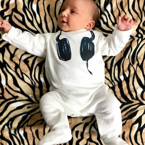 May include: A white long-sleeved footed sleeper for an infant featuring a graphic print of dark blue headphones on the chest. The garment has a round neckline and covers the feet, providing full coverage for the arms and legs in a solid white color.