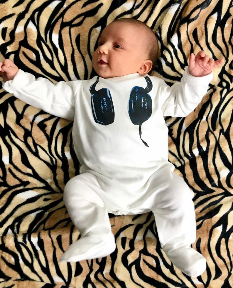 May include: A white long-sleeved footed sleeper for an infant featuring a graphic print of dark blue headphones on the chest. The garment has a round neckline and covers the feet, providing full coverage for the arms and legs in a solid white color.