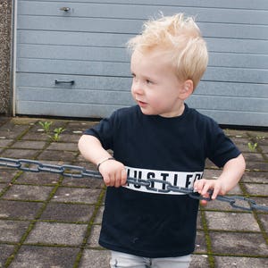 May include: A young boy wearing a black t-shirt with the word "HUSTLE" printed in white across the chest. He is holding a chain link fence in front of him and is looking to the left.