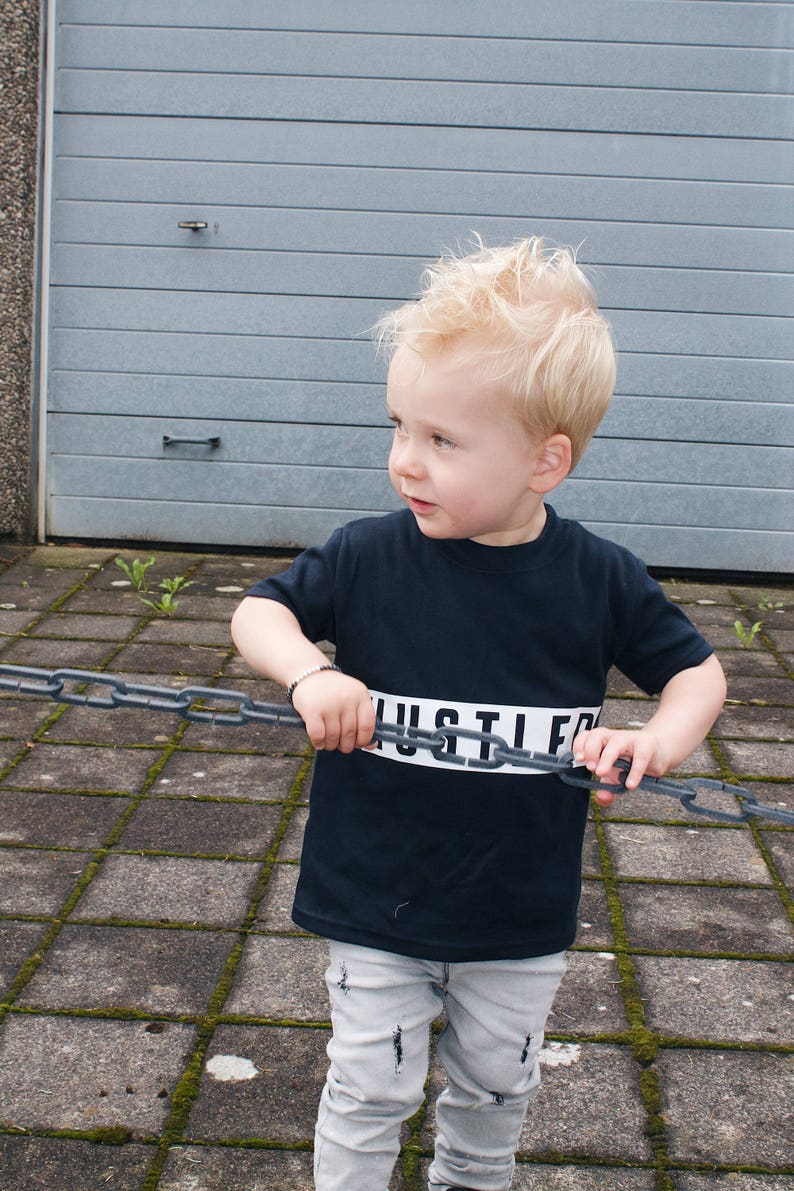 May include: A young boy wearing a black t-shirt with the word "HUSTLE" printed in white across the chest. He is holding a chain link fence in front of him and is looking to the left.