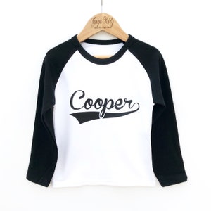 May include: White long-sleeve baseball tee with black sleeves. The shirt has a black graphic with the name "Cooper" in a vintage baseball font.