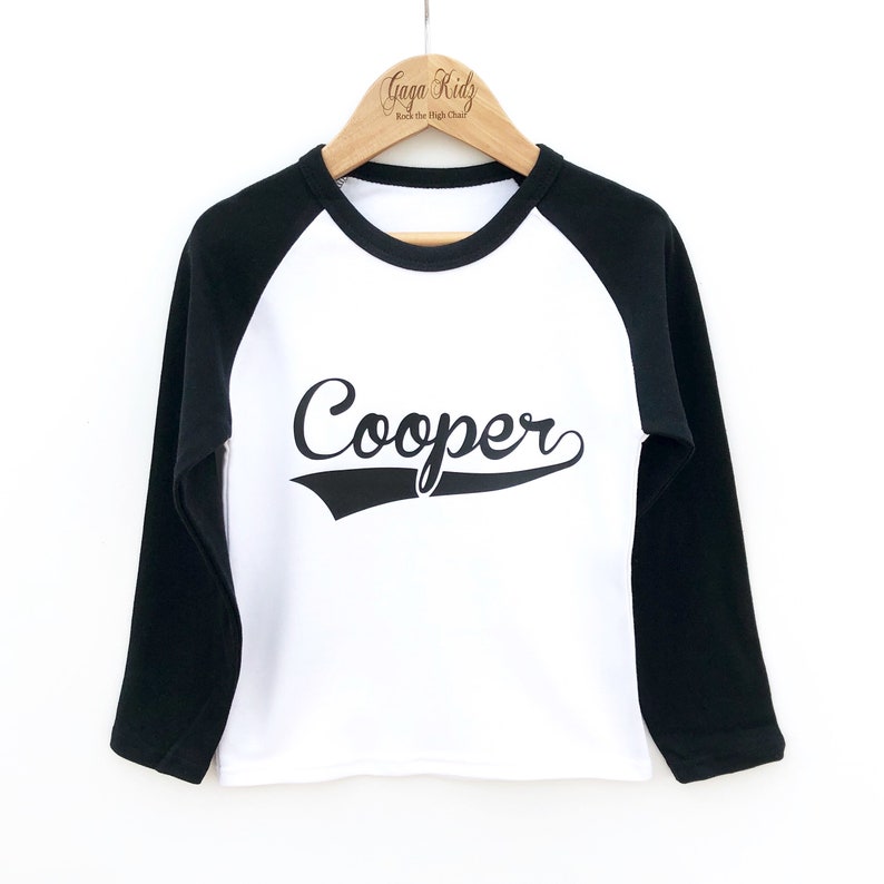 May include: White long-sleeve baseball tee with black sleeves. The shirt has a black graphic with the name "Cooper" in a vintage baseball font.