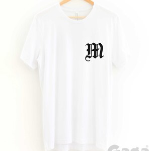 K&ouml;nnte beinhalten: Wei&szlig;es T-Shirt mit einem schwarzen "M" in gotischer Schrift auf der Vorderseite. Das Shirt h&auml;ngt an einem Holzb&uuml;gel.