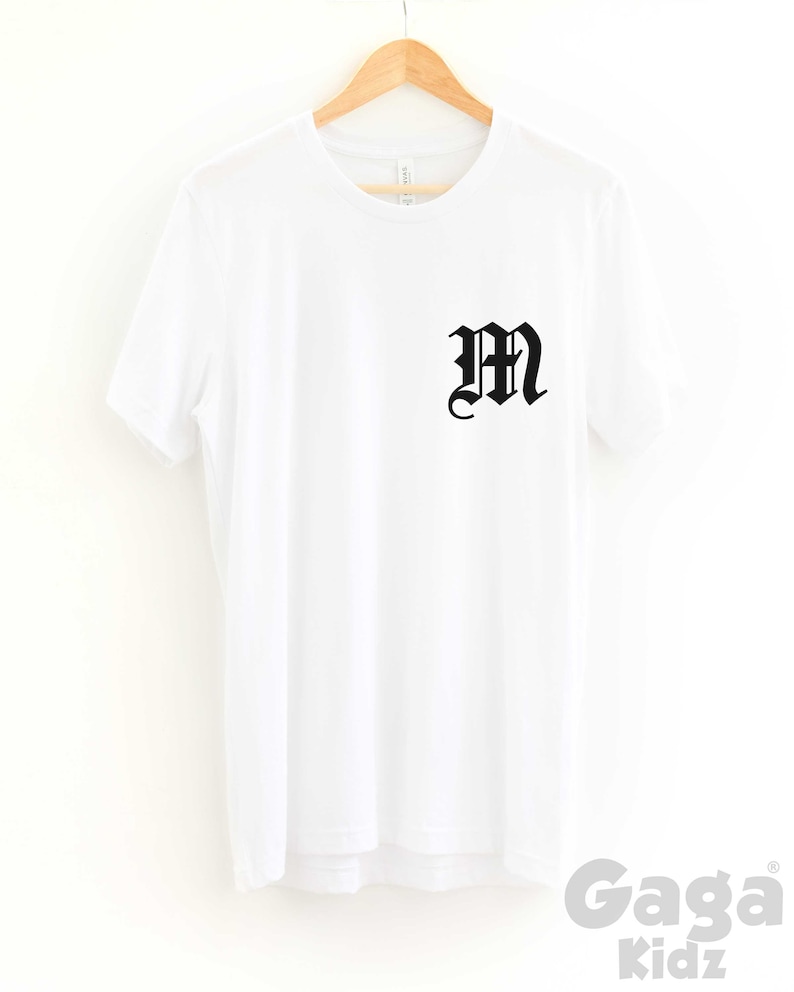 K&ouml;nnte beinhalten: Wei&szlig;es T-Shirt mit einem schwarzen "M" in gotischer Schrift auf der Vorderseite. Das Shirt h&auml;ngt an einem Holzb&uuml;gel.