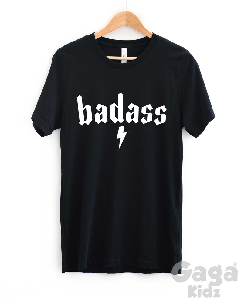 Może przedstawiać: Czarny T-shirt z białym napisem "badass" w stylu gotyckim z błyskawicą pod spodem.