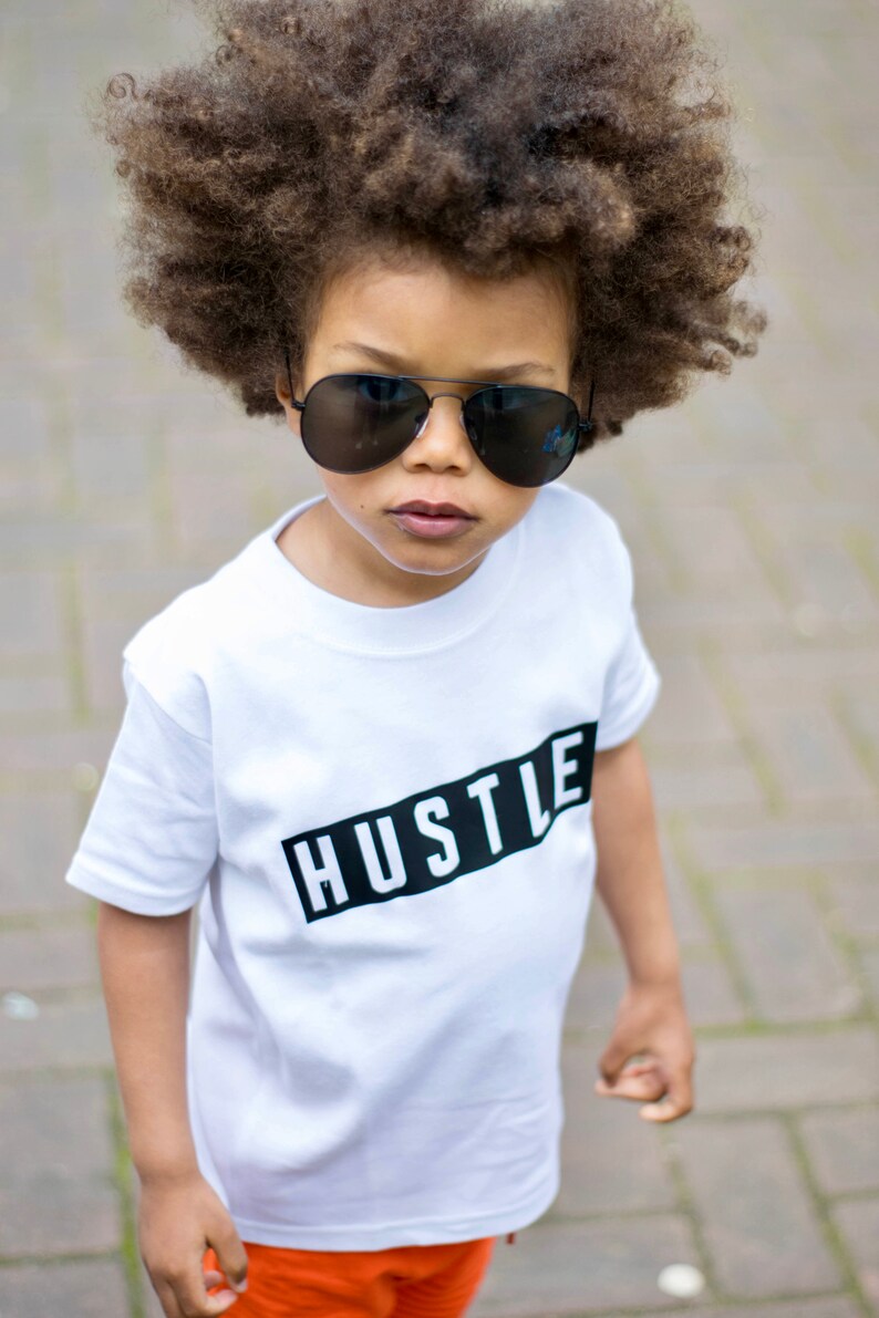 May include: A young boy with an afro hairstyle wears black sunglasses and a white t-shirt with the word "Hustle" printed in black on a black rectangle.
