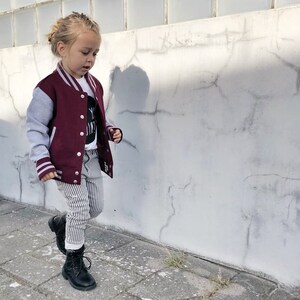 Op de afbeelding: Een jong kind draagt een bordeauxrode en grijze varsityjas met witte strepen, een gestreepte broek en zwarte laarzen. Het kind loopt naar een witte muur.