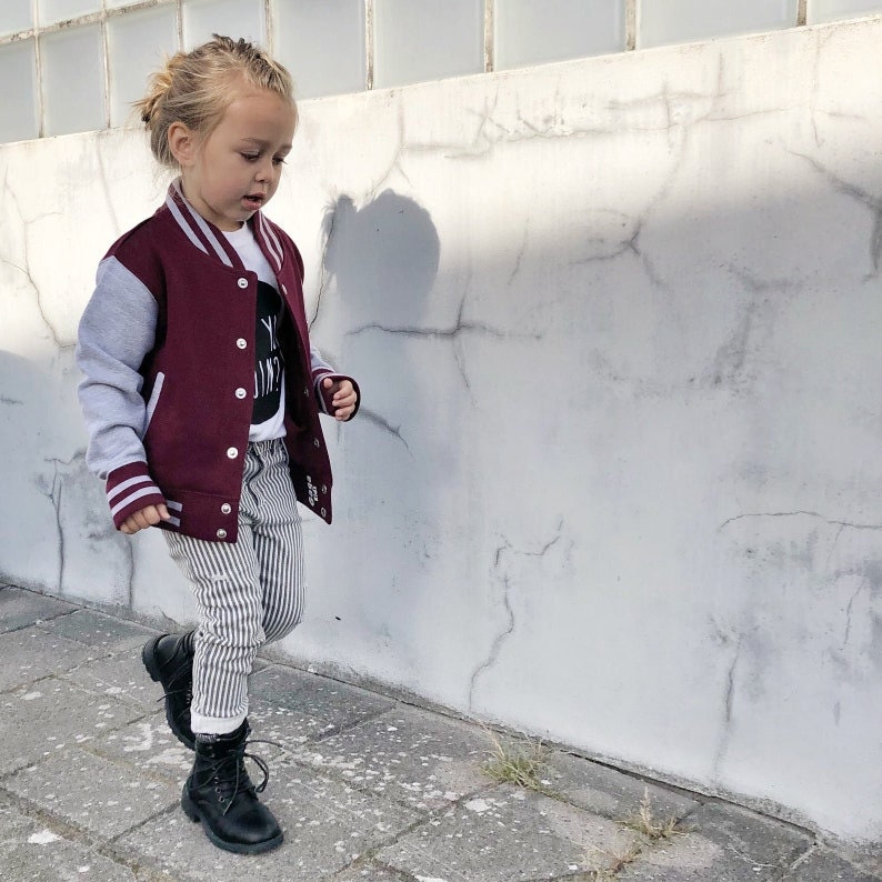 Op de afbeelding: Een jong kind draagt een bordeauxrode en grijze varsityjas met witte strepen, een gestreepte broek en zwarte laarzen. Het kind loopt naar een witte muur.