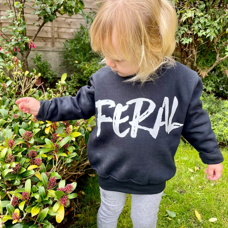 May include: A child wearing a black sweatshirt with the word "FERAL" printed in white on the front. The sweatshirt has a relaxed fit and a crew neck.