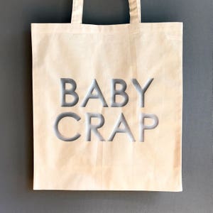 Pode incluir: Uma bolsa tote de lona branca com o texto "BABY CRAP" impresso em prata na frente.
