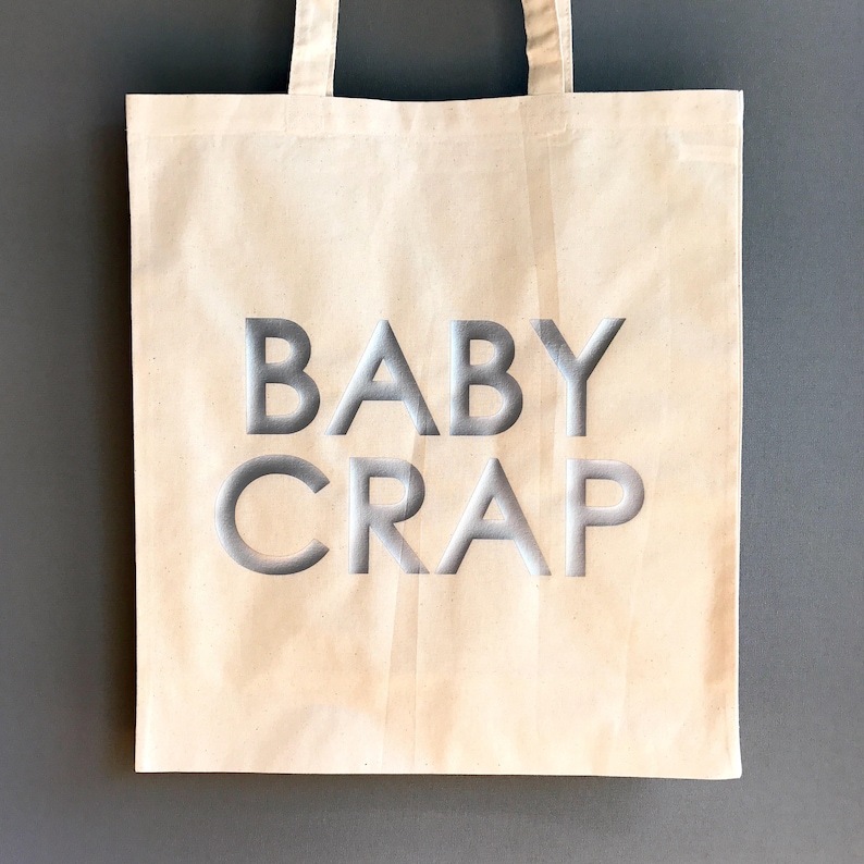 Pode incluir: Uma bolsa tote de lona branca com o texto "BABY CRAP" impresso em prata na frente.