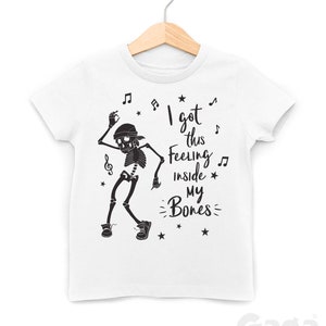 Peut inclure: T-shirt blanc avec un squelette noir dansant et le texte "I got this feeling inside my bones" avec des notes de musique et des &eacute;toiles.