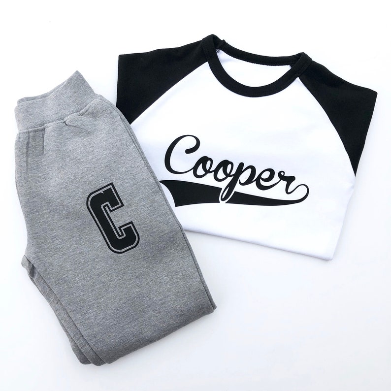 May include: A gray and black two-piece outfit for a child. The top is a white t-shirt with black sleeves and the word "Cooper" printed in black. The bottom is gray sweatpants with a black "C" printed on the left leg.