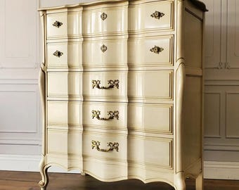 May include: A cream-colored, five-drawer chest of drawers with a glossy finish. The antique-style dresser features ornate brass hardware and curved legs. The drawers have a wavy design, adding to its vintage appeal.