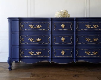 May include: A navy blue dresser with gold accents. The dresser has nine drawers with ornate gold hardware and trim. The piece has a curved design and sits on curved legs. A bouquet of white flowers rests on top.