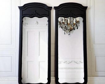 May include: Two tall, black framed mirrors with ornate tops. One mirror reflects a white door, the other a crystal chandelier. The mirrors stand on a wooden floor against a white paneled wall, creating a classic, elegant aesthetic.