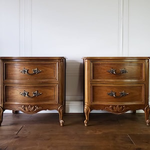May include: Two matching wooden nightstands with two drawers each. The nightstands have ornate brass handles and decorative carvings on the lower portion. The wood has a warm, medium brown tone. The nightstands are on a wooden floor.
