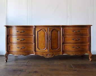May include: A wooden dresser with a warm brown finish. It features six drawers on the sides and two doors in the center, all adorned with ornate hardware. The dresser has curved legs and decorative carvings.