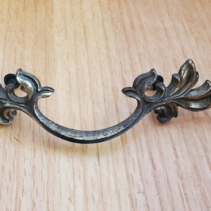 May include: Antique bronze-toned drawer pull with a curved handle and ornate leaf design. The decorative hardware is made of metal and is suitable for cabinets, drawers, or furniture. The pull measures approximately 6 inches in length.