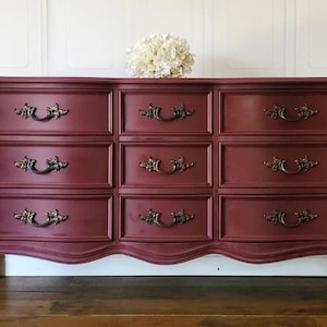 May include: A burgundy dresser with nine drawers and ornate brass handles. The dresser has a curved base and decorative legs. A white floral arrangement sits on top of the dresser.