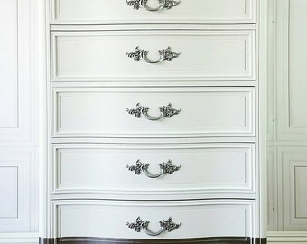 May include: A white six-drawer chest of drawers with ornate silver hardware. The bottom of the chest and the legs are silver. The top of the chest has a curved edge. The chest is decorated with white flowers.