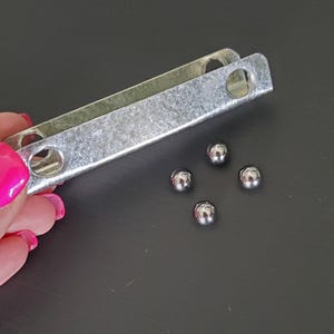 May include: A close-up of a metal bracket with four small, shiny, silver metal balls. The bracket is rectangular with rounded corners and holes. The background is a dark gray surface.