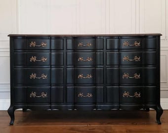 May include: A black dresser with nine drawers and ornate gold-colored handles. The top is a dark wood, and the dresser has curved legs. The drawers are arranged in a grid pattern.
