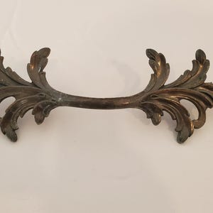 May include: Ornate bronze-toned cabinet pull with a symmetrical design. The pull features a curved bar with stylized leaf and scroll details on each end. The hardware is likely for a drawer or cabinet door.
