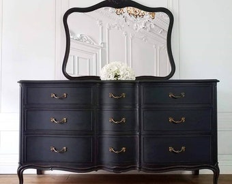 May include: A black dresser with nine drawers and ornate brass handles. A matching black framed mirror with a decorative top is above the dresser. A bouquet of white flowers sits on the dresser. The dresser has curved legs and a curved bottom.